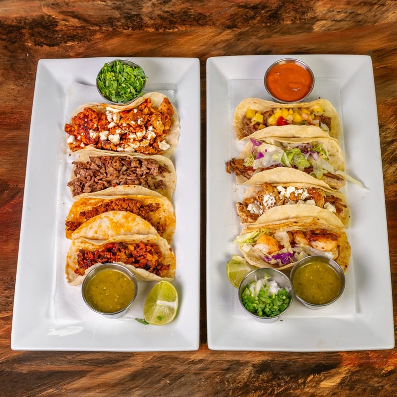 Two white plates filled with various MexCantina tacos, salsas, and lime wedges on a wooden table.