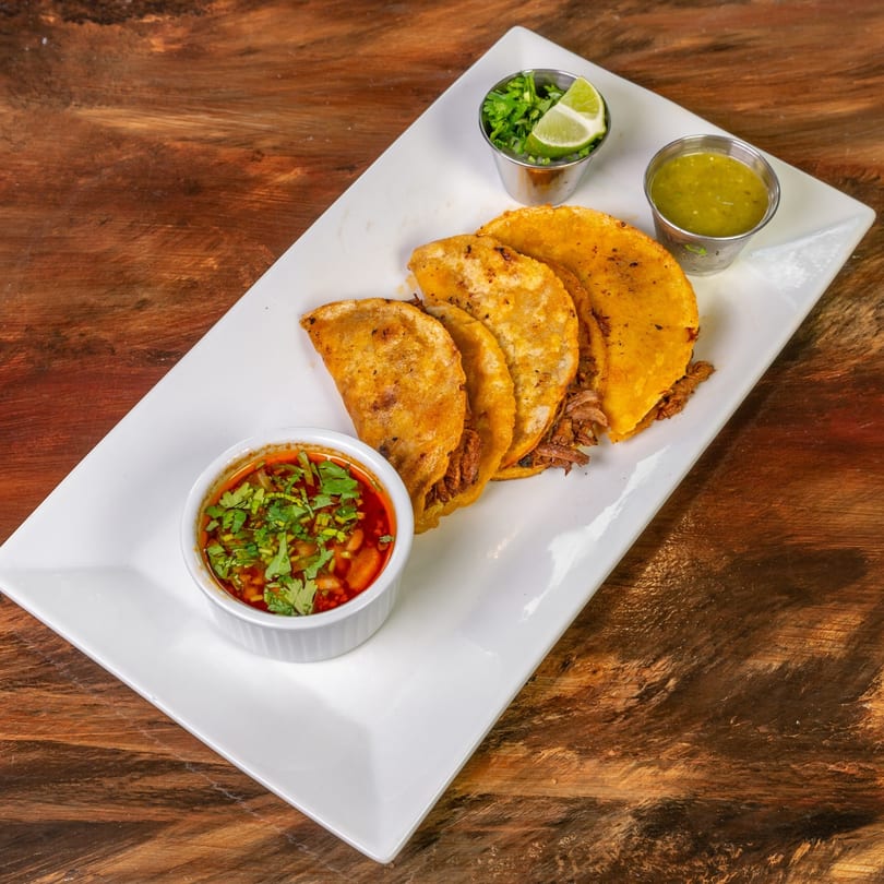 Four MexCantina birria tacos with consome, green salsa, cilantro, and lime on a white plate.